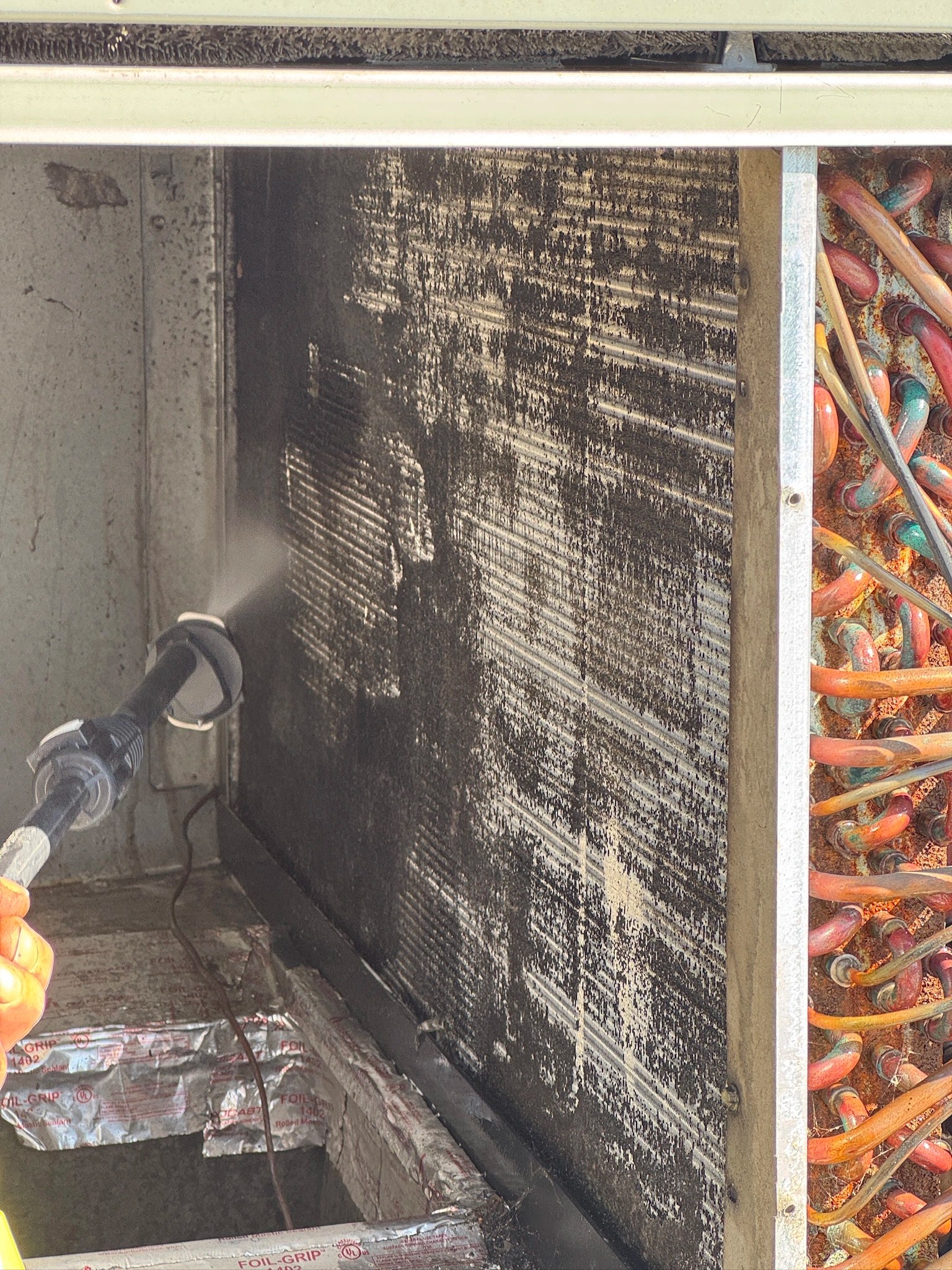 Technician pressure washing a dirty evaporator coil inside a commercial HVAC unit, revealing clean fins underneath